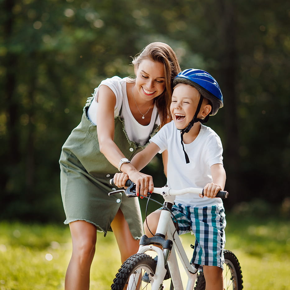 Smiling woman helping a boy wearing a helmet ride a bicycle in a park.